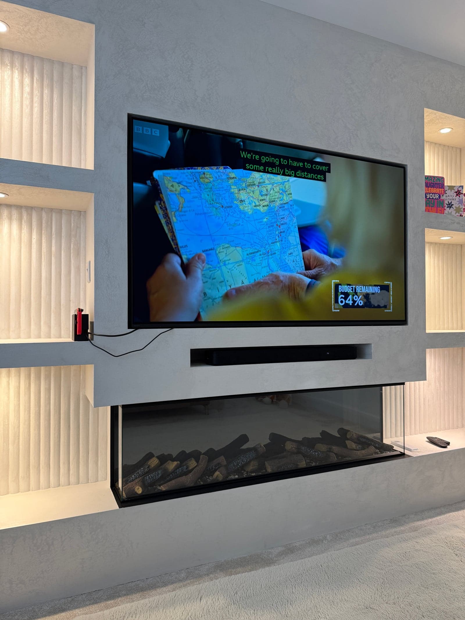 Recessed wall-mounted TV on a grey microcement media wall with fluted LED-lit niches, a concealed soundbar and an inset log-effect electric fire below