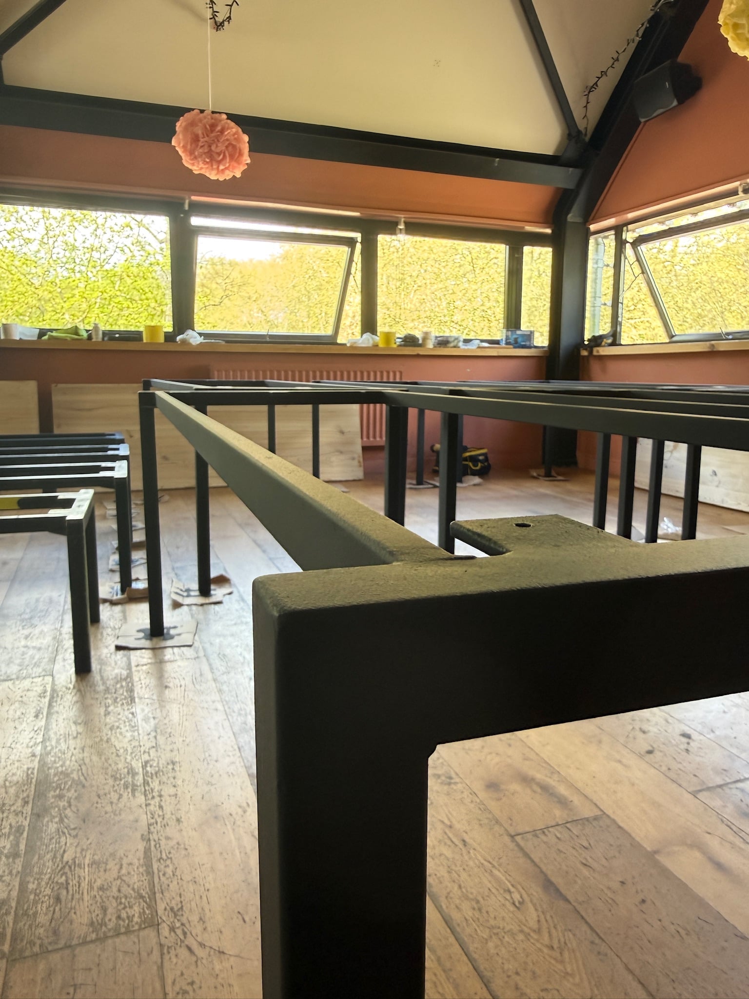 Commercial cafe interior with black powder-coated steel table frames awaiting tops, oak flooring and warm terracotta walls